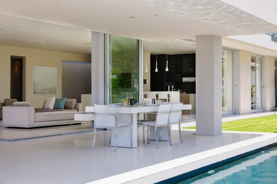 Modern coastal living room with slide-away glass doors opening to a patio and pool, creating a seamless indoor-outdoor living space with natural light and outdoor dining seating.