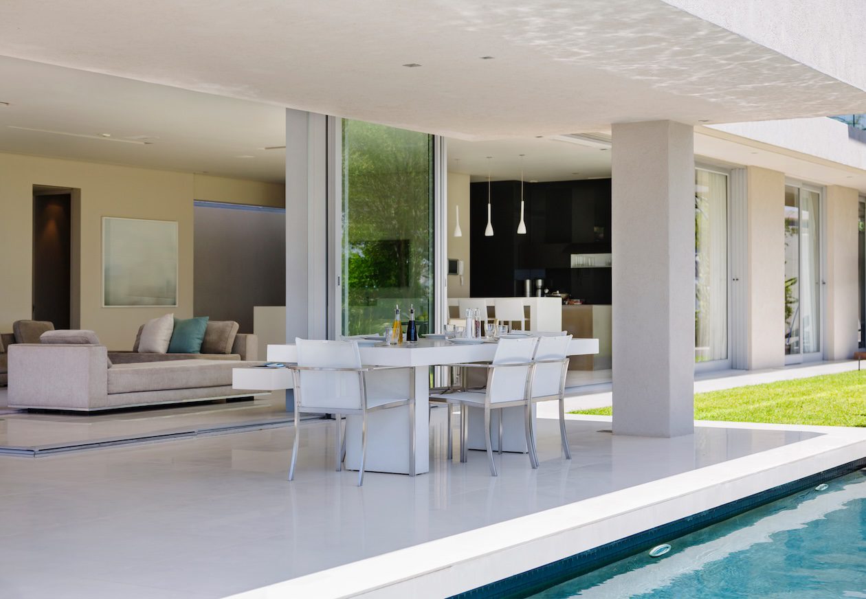Modern coastal living room with slide-away glass doors opening to a patio and pool, creating a seamless indoor-outdoor living space with natural light and outdoor dining seating.