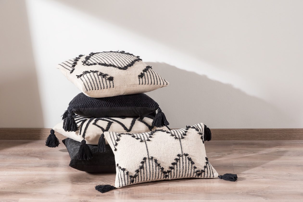 Stacked decorative throw pillows in black and cream patterns with tassels, including square and lumbar pillow sizes styled on a wood floor against a neutral wall