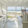 Modern coastal-style living room viewed from outside through large glass windows, featuring neutral furniture, layered textures, natural light, and an open staircase in a contemporary California casual home.