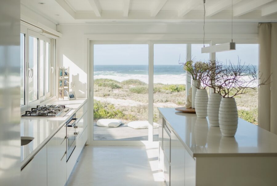 Modern coastal-style living room viewed from outside through large glass windows, featuring neutral furniture, layered textures, natural light, and an open staircase in a contemporary California casual home.