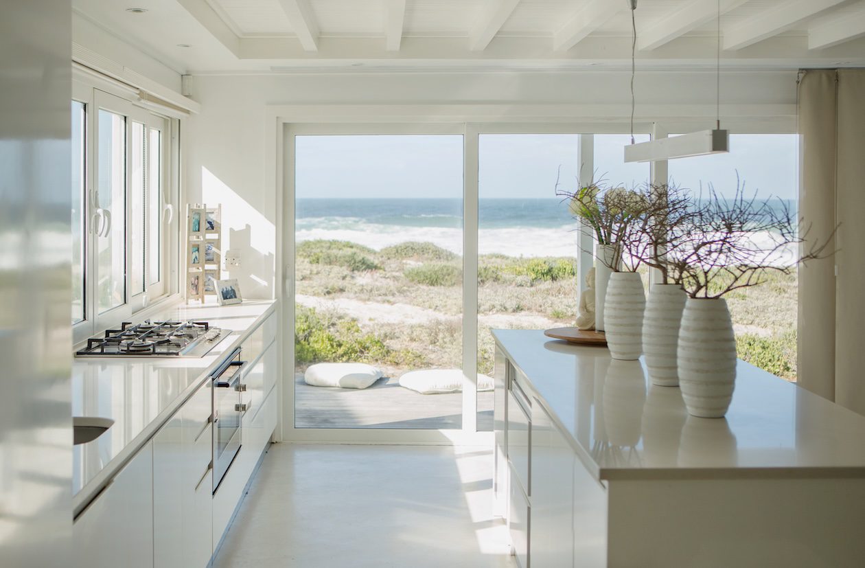 Modern coastal-style living room viewed from outside through large glass windows, featuring neutral furniture, layered textures, natural light, and an open staircase in a contemporary California casual home.