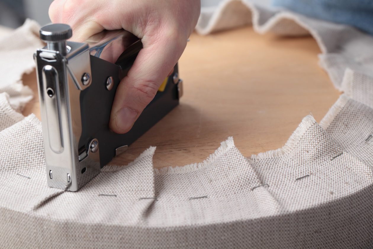 Hand using an upholstery staple gun to attach fabric to a wooden furniture frame during the custom furniture reupholstery process.