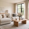 California casual living room with warm wood furniture, neutral upholstery, woven textures, and large windows overlooking greenery.